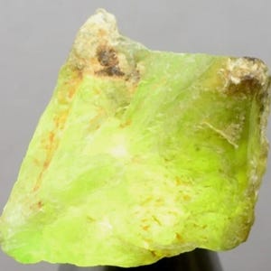 May include: A raw, translucent mineral specimen featuring a vibrant lime-green color. The stone has an irregular, jagged shape with visible crystalline textures, internal fractures, and small patches of brownish-orange surface inclusions.