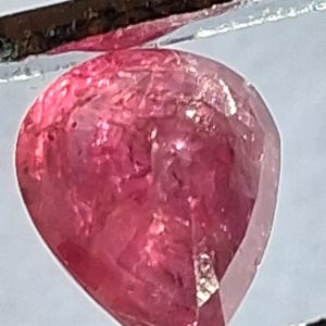 May include: A pear-shaped, faceted gemstone with a vibrant, translucent pink hue. The stone displays internal light reflections and a polished surface, held securely between two dark, thin metal prongs against a light background.