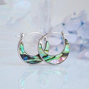 May include: A pair of silver hoop earrings with a crescent-shaped abalone shell inlay. The shell has iridescent green, blue, and purple colors.
