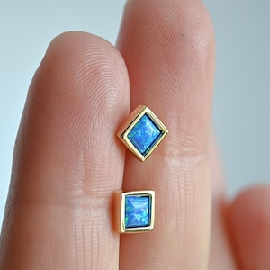 May include: Two gold-colored square stud earrings with a blue opal center. The earrings are held between two fingers. The opal has a shimmering, iridescent quality. The earrings have a simple, elegant design.