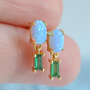 May include: A pair of gold earrings with light blue opal stones and green emeralds. The earrings are held up by a hand. The opal stones are oval-shaped, and the emeralds are rectangular.