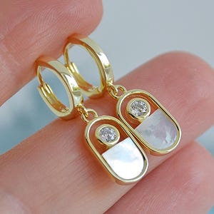 May include: Gold-tone hoop earrings with a hanging oval pendant. Each pendant has a white, iridescent inlay and a small, clear gemstone. The earrings are held by a hand against a soft, blurred background.