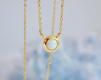 May include: A delicate gold necklace featuring a round pendant with a white opal stone. The pendant is set in a gold bezel and hangs from a fine gold chain. The necklace is displayed against a soft, blurred background.
