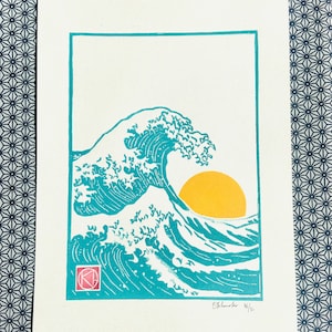 May include: A print of a turquoise wave with a yellow sun in the background. The artwork is framed in turquoise and includes the artist's signature and edition number. The print is on white paper.