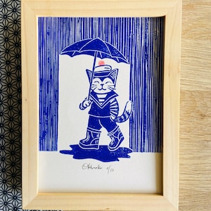 Op de afbeelding: Blauw-witte prent van een kat in matrozenkleding die in de regen loopt onder een paraplu. Het kunstwerk is ingelijst in een licht houten frame. De prent is genummerd 1/50 en gesigneerd.