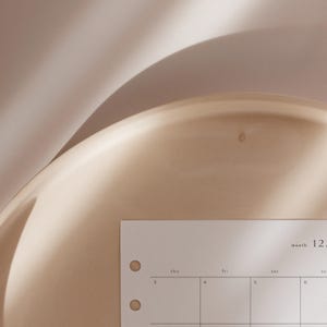 K&ouml;nnte beinhalten: Nahaufnahme eines beigefarbenen Keramiktabletts mit einer Kalenderseite. Die Kalenderseite ist wei&szlig; mit schwarzem Text und zeigt den Monat und das Datum. Das Tablett hat eine glatte, gl&auml;nzende Oberfl&auml;che.