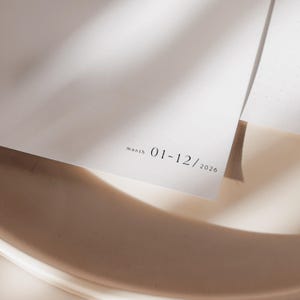 K&ouml;nnte beinhalten: Nahaufnahme eines wei&szlig;en Papiers mit dem Text "month 01-12/2026" in einer minimalistischen Schriftart. Das Papier ist leicht angewinkelt, mit weichen Schatten und einem neutralen Hintergrund. Das Bild deutet auf einen Kalender oder Planer hin.