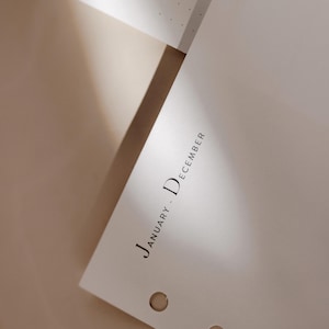 K&ouml;nnte beinhalten: Nahaufnahme einer wei&szlig;en Planerseite mit dem Text "January - December" in minimalistischer schwarzer Schrift. Die Seite hat drei Bindungsl&ouml;cher und ist vor einem neutralen Hintergrund abgewinkelt.