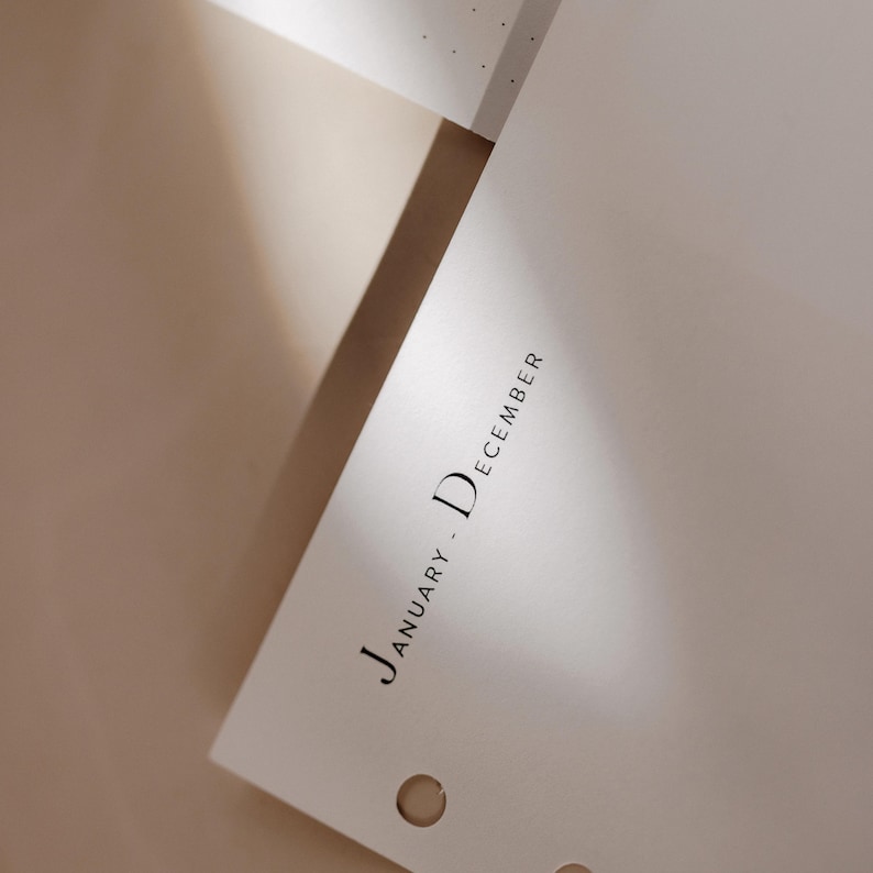K&ouml;nnte beinhalten: Nahaufnahme einer wei&szlig;en Planerseite mit dem Text "January - December" in minimalistischer schwarzer Schrift. Die Seite hat drei Bindungsl&ouml;cher und ist vor einem neutralen Hintergrund abgewinkelt.