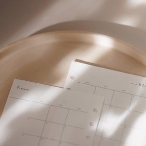 K&ouml;nnte beinhalten: Nahaufnahme eines beigefarbenen Kalenders mit dem Monat Februar. Die Kalenderseiten sind hell und haben ein Rasterlayout. Der Kalender liegt auf einer beigefarbenen Oberfl&auml;che, wobei weiche Schatten Tiefe verleihen. Die Gesamt&auml;sthetik ist minimalistisch und neutral.