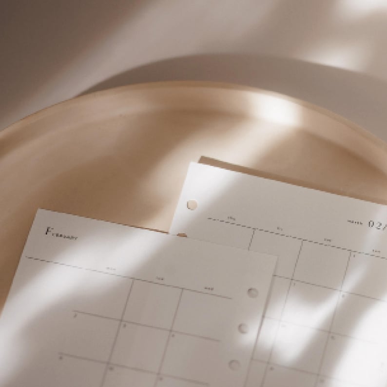 K&ouml;nnte beinhalten: Nahaufnahme eines beigefarbenen Kalenders mit dem Monat Februar. Die Kalenderseiten sind hell und haben ein Rasterlayout. Der Kalender liegt auf einer beigefarbenen Oberfl&auml;che, wobei weiche Schatten Tiefe verleihen. Die Gesamt&auml;sthetik ist minimalistisch und neutral.