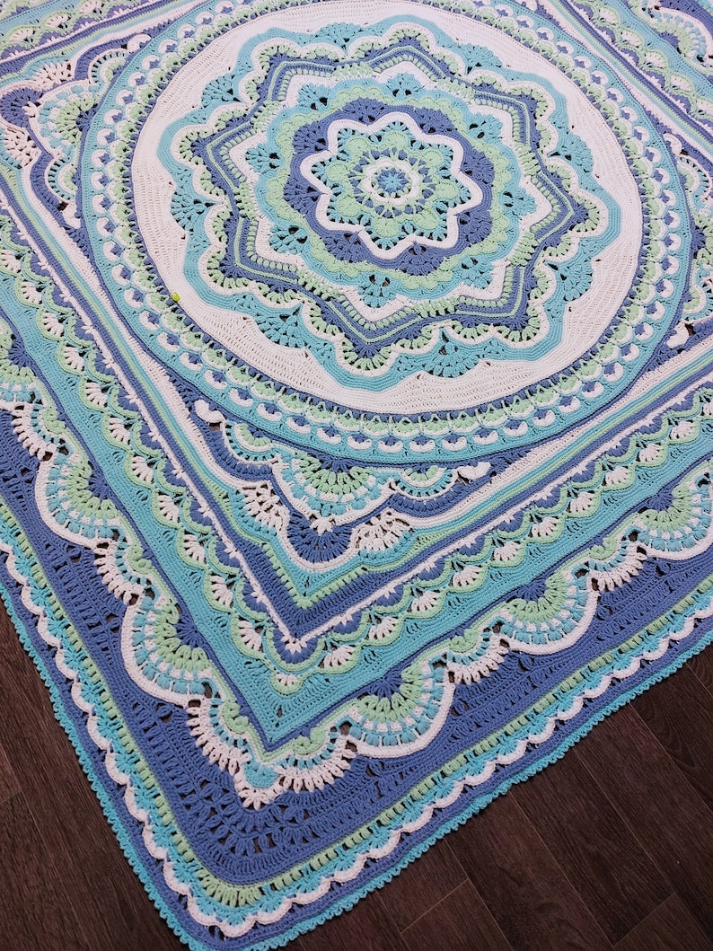 May include: A large, square, crocheted blanket in shades of blue, teal, and white. The intricate design features a central floral motif surrounded by concentric circles and scalloped edges. The blanket is made of soft yarn and is perfect for a bed or sofa.