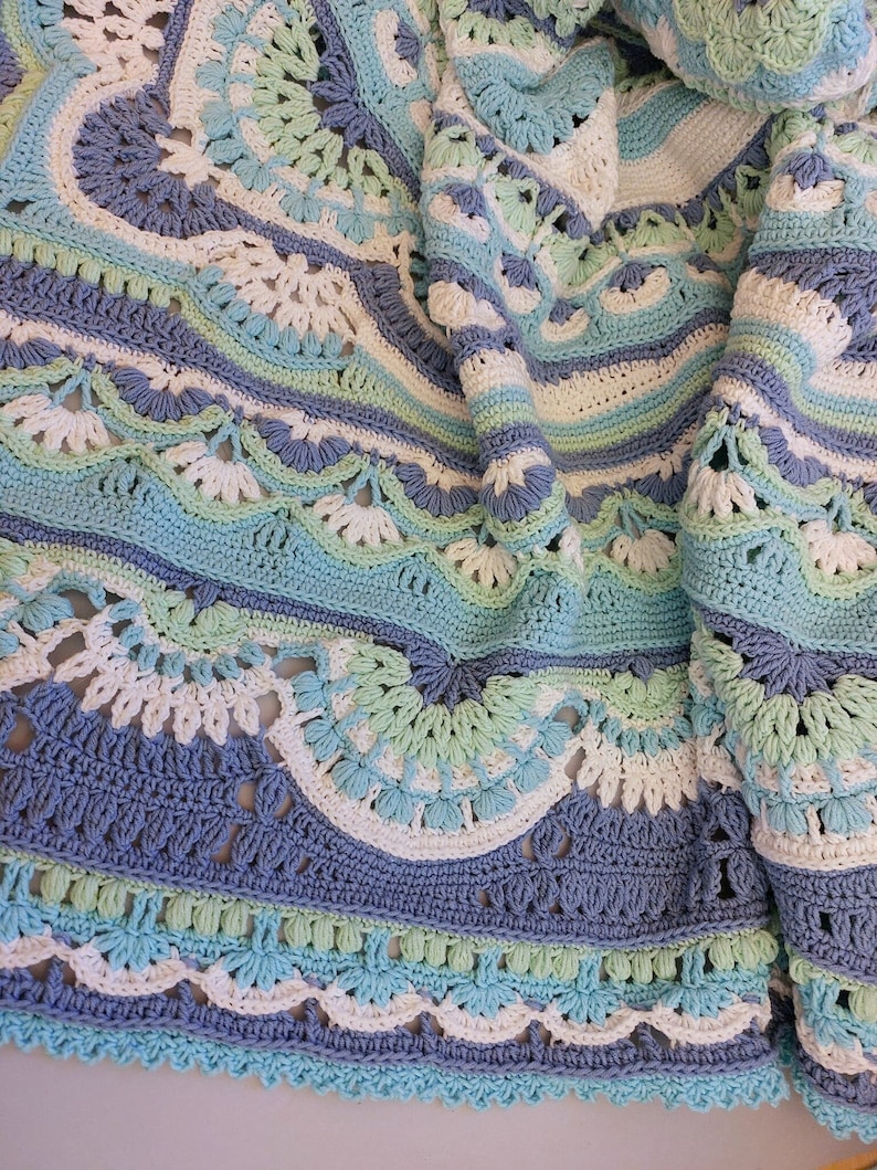May include: A handmade crochet blanket in shades of blue, white, and green. The blanket features intricate circular patterns and scalloped edges. The soft color palette and detailed stitching create a cozy, decorative textile.