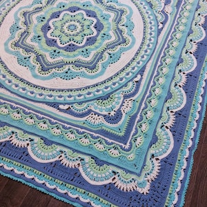 May include: A large, square, crocheted blanket in shades of blue, white, and green. The design features a central floral motif surrounded by concentric geometric patterns and scalloped edges. The blanket is handmade.