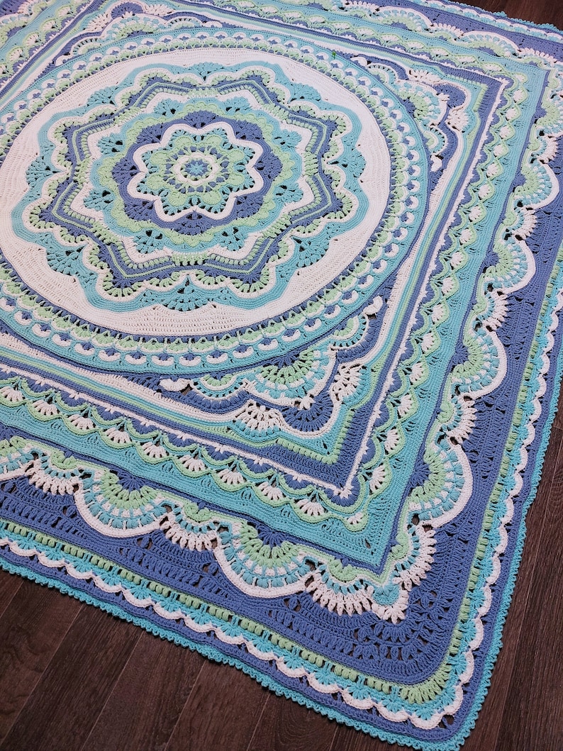 May include: A large, square, crocheted blanket in shades of blue, white, and green. The design features a central floral motif surrounded by concentric geometric patterns and scalloped edges. The blanket is handmade.