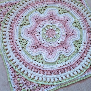 Pode incluir: Uma manta quadrada de crochê com um padrão de mandala circular no centro. O design incorpora anéis concêntricos de fio rosa suave, verde sálvia e branco, utilizando um trabalho de pontos intrincado que inclui pontos pipoca e conchas.