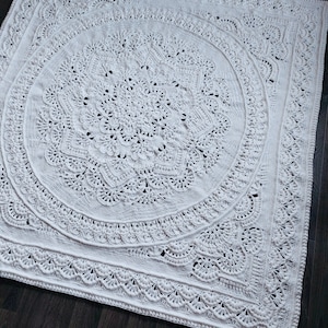 May include: A large, off-white crochet blanket with an intricate, circular mandala design. The blanket features detailed patterns and a scalloped edge. The handmade blanket is displayed on a dark wood surface.