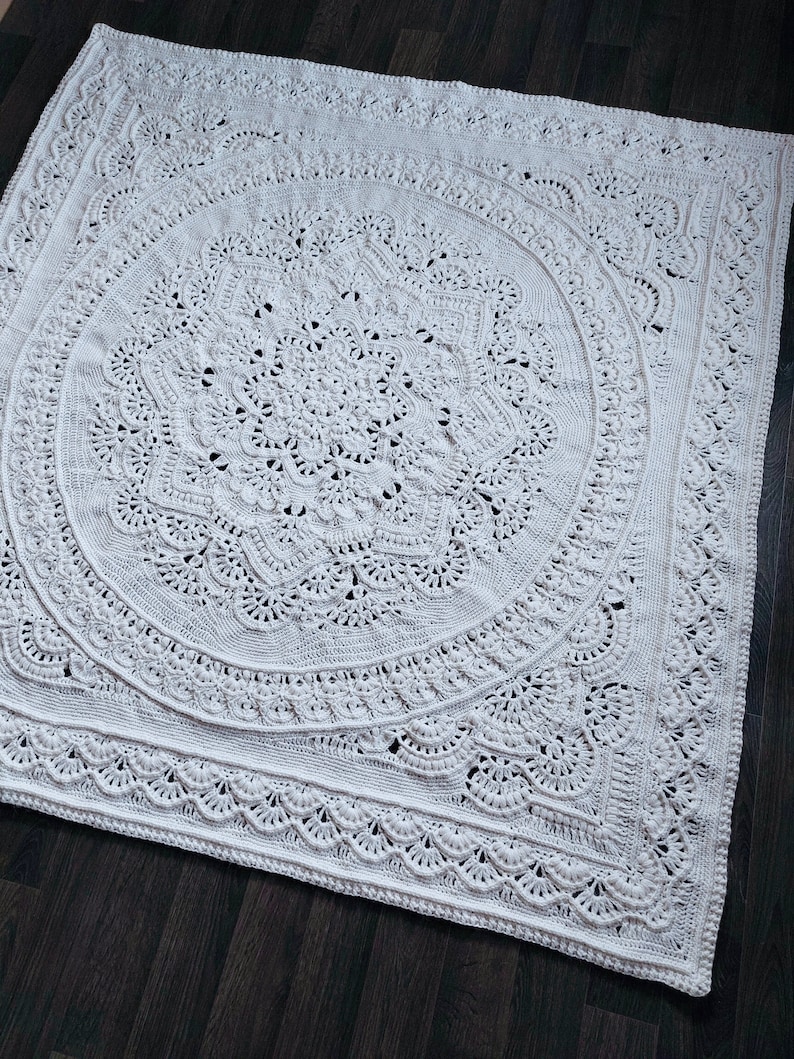 May include: A large, off-white crochet blanket with an intricate, circular mandala design. The blanket features detailed patterns and a scalloped edge. The handmade blanket is displayed on a dark wood surface.