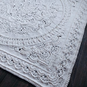 May include: A large, off-white, crocheted blanket with an intricate, circular design. The blanket features detailed patterns, including floral and scalloped edges. The texture is visible, showcasing the handmade quality of the piece. The blanket is placed on a dark wooden surface.