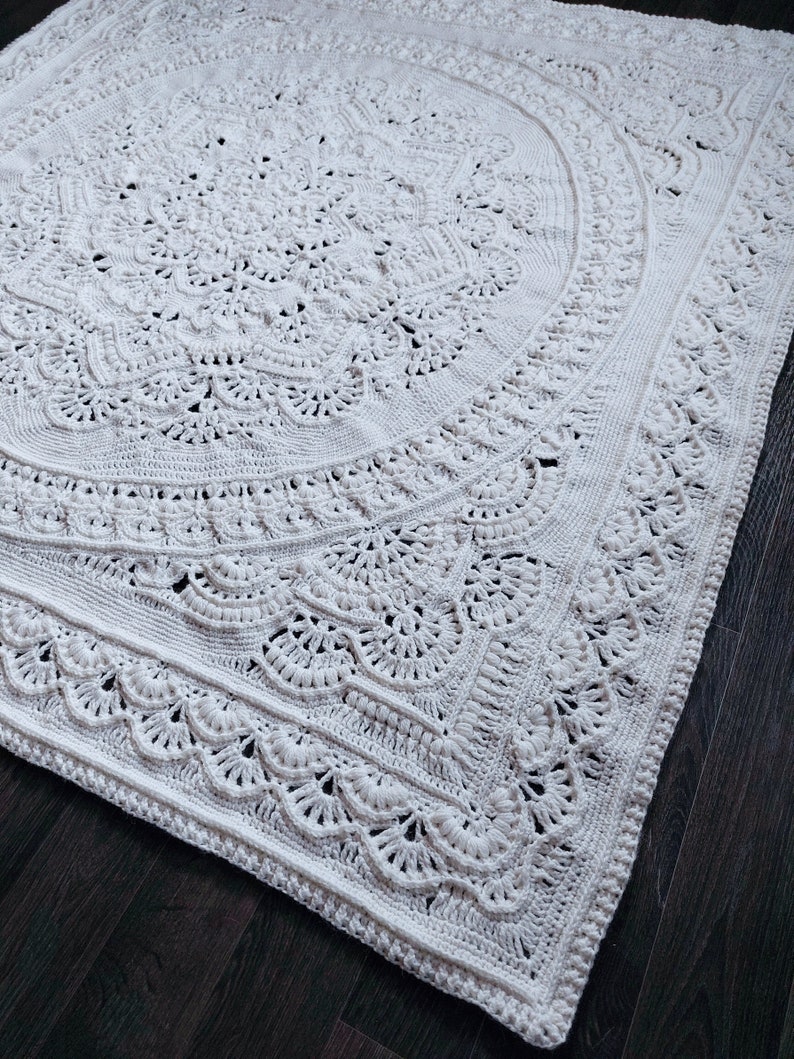 May include: A large, off-white, crocheted blanket with an intricate, circular design. The blanket features detailed patterns, including floral and scalloped edges. The texture is visible, showcasing the handmade quality of the piece. The blanket is placed on a dark wooden surface.