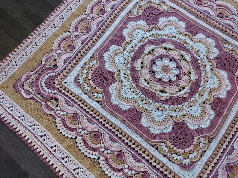 Peut inclure: Une couverture en crochet avec un grand motif de mandala complexe dans des tons de rose, blanc et marron. La couverture a une bordure de motifs plus petits et festonn&eacute;s.