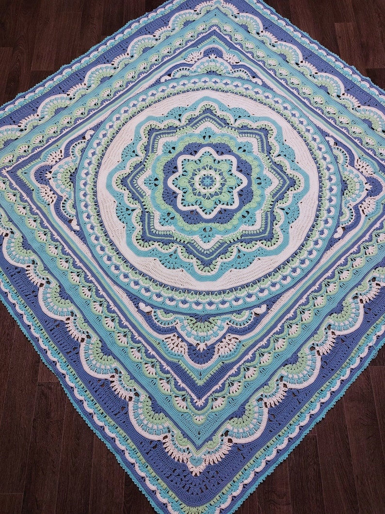 May include: A handcrafted, square crochet blanket in shades of blue, white, and green. The intricate design features a central floral motif surrounded by concentric circles and scalloped edges. The blanket is made with soft yarn.