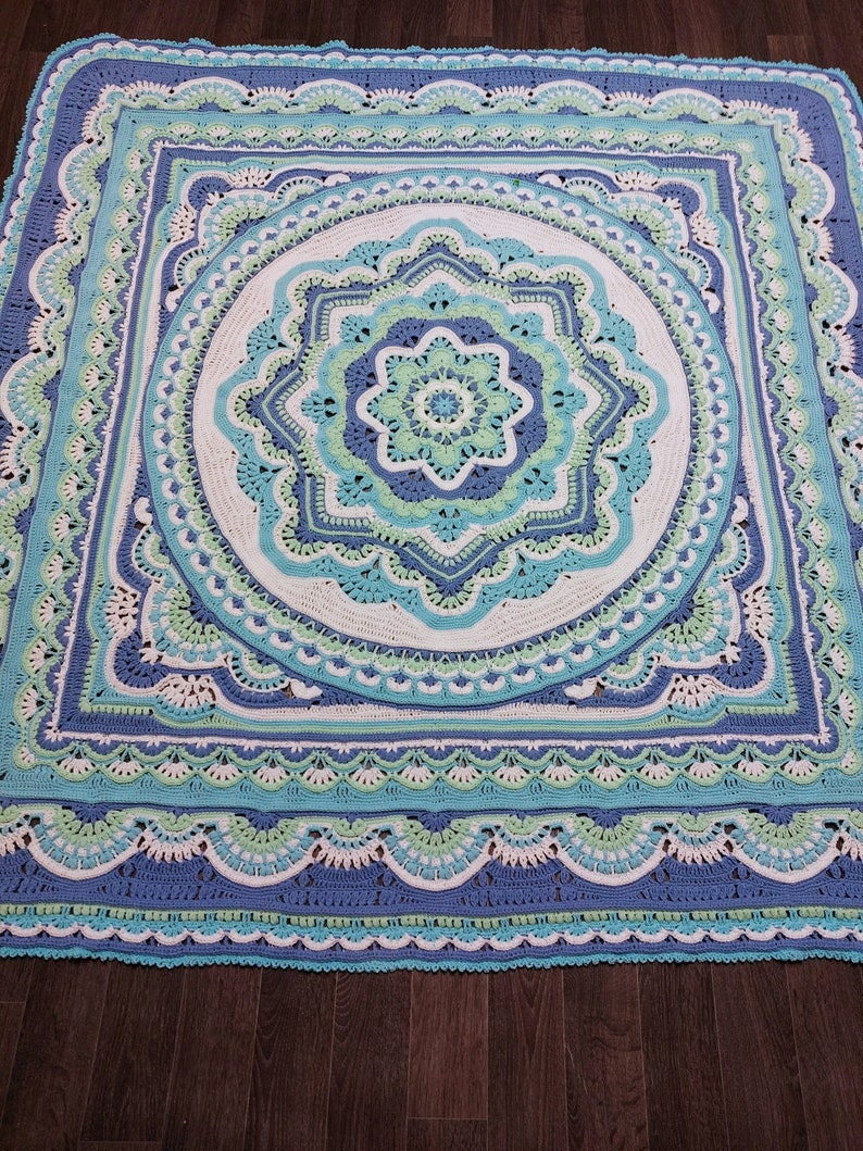 May include: A large, square, crocheted blanket in shades of blue, white, and green. The blanket features a detailed, circular mandala design in the center, surrounded by intricate patterns and scalloped edges. The color palette includes light and dark blues, white, and a touch of green.