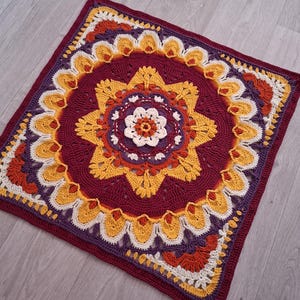 May include: A square crochet blanket featuring a central white flower surrounded by concentric layers of yellow, orange, and deep burgundy yarn. The intricate design includes petal-like motifs and geometric patterns that radiate outward to the edges.