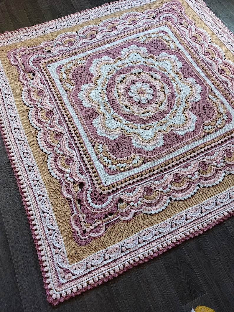 Peut inclure: Une couverture au crochet avec un grand motif floral complexe dans des tons de rose, blanc et beige. La couverture a une bordure festonn&eacute;e et est r&eacute;alis&eacute;e avec une vari&eacute;t&eacute; de points.