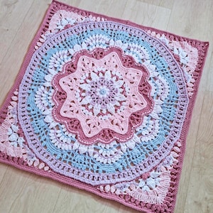 May include: A square crochet blanket featuring a central mandala design with concentric layers in shades of dusty rose, light pink, white, and pale blue. The intricate pattern transitions from a circular floral centre to a square border.