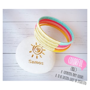 Könnte beinhalten: Ein Set aus bunten Armbändern mit einem goldenen Glitzerakzent. Die Armbänder sind übereinander gestapelt und in einem Kreis angeordnet. Die Armbänder befinden sich auf einer weißen Oberfläche mit einer Sonne und dem Wort "Samos" darauf.