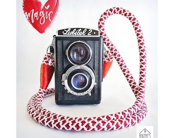 May include: A red and white camera strap with a black Lubitel 2 camera attached. The camera has two lenses and the strap is made of braided rope. The background is white with a red watercolor heart that says "magic" in white lettering.