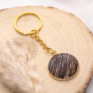 Könnte beinhalten: Ein goldfarbener Schlüsselanhänger mit einem kreisförmigen Anhänger. Der Anhänger hat ein braunes und weißes Muster, das in einem goldfarbenen Rahmen eingefasst ist. Der Schlüsselanhänger liegt auf einer Holzoberfläche.