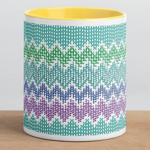 Puede incluir: Una taza de cerámica con interior amarillo brillante y exterior blanco que presenta un patrón horizontal repetitivo de chevrón en tonos azul verdoso, verde, azul y morado. El diseño imita una textura tejida con detalles de punto de cruz.