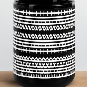 Puede incluir: Una taza de cerámica negra con un patrón geométrico blanco repetitivo que envuelve el exterior. El diseño consta de bandas horizontales de varias formas, incluyendo líneas, guiones, zigzags y pequeños triángulos en un marcado contraste.