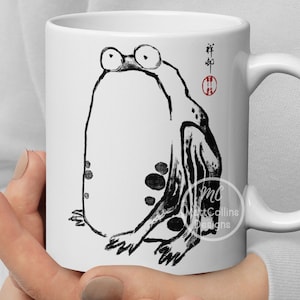 Może przedstawiać: Biały kubek ceramiczny z czarno-białym rysunkiem żaby po jednej stronie. Żaba siedzi ze skrzyżowanymi nogami i ma duży, okrągły brzuch. Kubek ma czarny uchwyt, a na spodzie nadrukowany jest napis "Art Collins Designs".