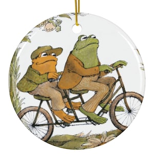 Op de afbeelding: Ronde keramische kerstversiering met een illustratie van twee kikkers op een tandemfiets. Eén kikker draagt een groene jas, de ander een bruine jas. De versiering heeft een gouden touwtje om op te hangen. Diameter: ongeveer 7,6 cm.