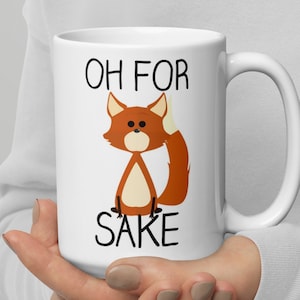 以下が含まれることがあります： 白いセラミック製のコーヒーカップに、漫画のキツネと「Oh for sake」という文字が書かれています。