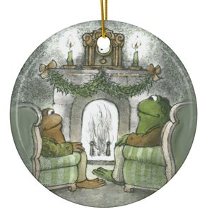 May include: Round ceramic ornament featuring a cozy scene with two frog characters seated in armchairs by a fireplace. The illustration has a vintage style with soft colors and a gold hanging string.