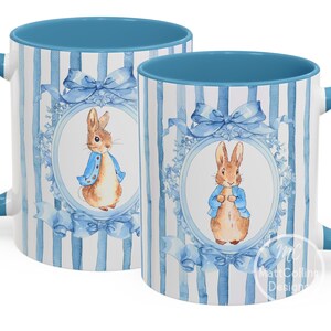 Puede incluir: Dos tazas de cerámica con asas e interiores azules. Las tazas presentan un patrón de rayas azules y blancas con una ilustración de acuarela de un conejo con una chaqueta azul. El diseño incluye lazos azules y detalles florales.