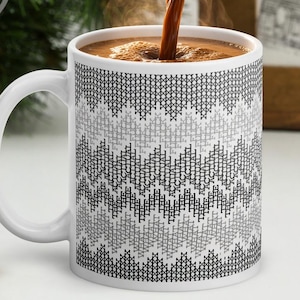 Puede incluir: Una taza de cerámica blanca con un patrón de chevrón repetitivo en negro y gris que imita una textura de punto de cruz. Se vierte líquido marrón oscuro en la taza, creando vapor. La taza tiene un asa curva y un acabado suave y brillante.
