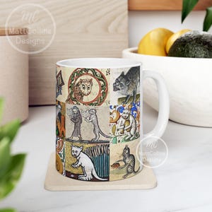 May include: White ceramic mug featuring a collage of medieval illustrations of animals and figures. The artwork includes owls, cats, and people reading books, set against a parchment-like background. The mug has a curved handle and is sitting on a beige coaster.