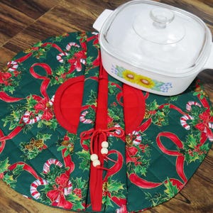 May include: A round, quilted fabric casserole carrier featuring a dark green background with a pattern of red ribbons, candy canes, and holly leaves, secured with red ties and wooden beads, alongside a white square glass baking dish with a lid.