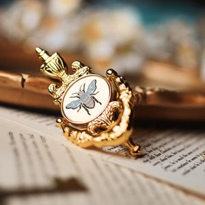 May include: A gold-toned door knocker with a round, white enamel centre featuring a detailed illustration of a bee. The knocker has a decorative, ornate design.