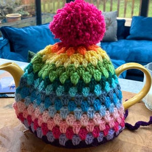 Peut inclure: Une théière en céramique jaune recouverte d'une housse au crochet faite à la main, présentant un motif arc-en-ciel dégradé de groupes de fils. La housse est surmontée d'un grand pompon en laine rose vif et d'un fil violet foncé à la base.