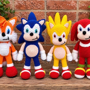 May include: Four handmade crochet plush toys representing characters from the Sonic the Hedgehog series. The figures include an orange fox with two tails, a blue hedgehog, a yellow hedgehog with spiked hair, and a red echidna with white gloves.