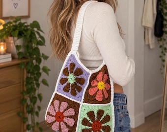 May include: A crochet tote bag constructed from four square panels, each featuring a large flower motif in shades of brown, pink, peach, and light green against lavender and white backgrounds, finished with a white crocheted shoulder strap.