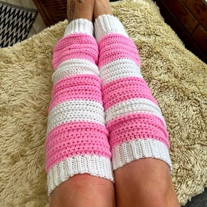 May include: A pair of leg warmers featuring a horizontal striped pattern in alternating bands of bright pink and white. The leg warmers are constructed with a textured crochet stitch and finished with ribbed white cuffs at the top and bottom edges.
