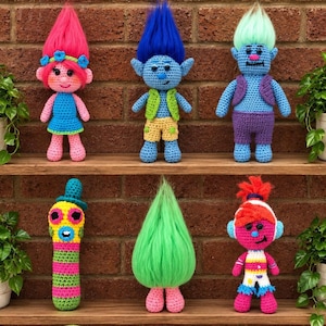 Könnte beinhalten: Eine Sammlung von sechs gehäkelten Puppen mit verschiedenen Designs, darunter trollartige Figuren mit leuchtend pinkem, blauem, grünem und orangefarbenem Haar sowie ein bunt gemustertes Wesen, die alle auf zwei Holzregalen präsentiert werden.