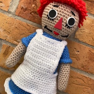 May include: A crocheted rag doll with red yarn hair, a white apron, and a blue dress. The doll has black button eyes and a red felt nose.